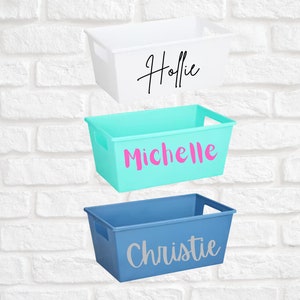 May include: Three rectangular storage bins in white, teal, and blue are displayed against a white brick wall. Each bin has a name printed on the front: "Hollie" in black, "Michelle" in pink, and "Christie" in silver. The bins have built-in handles.
