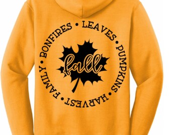 May include: A mustard yellow hooded sweatshirt featuring a black graphic on the back. The design includes a central maple leaf with the word fall in script, surrounded by a circular border of the words bonfires, leaves, pumpkins, harvest, and family.