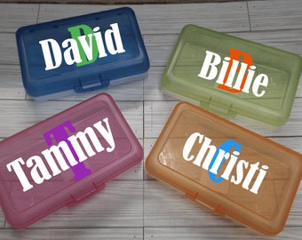 May include: Four personalized pencil boxes in blue, green, pink, and orange. Each box has a name in white lettering with a colored initial. The names are David, Billie, Tammy, and Christi.