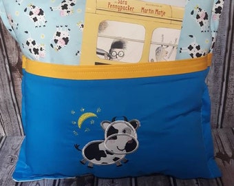 Pode incluir: Uma almofada azul com um bolso amarelo e um desenho de vaca. A almofada tem um topo azul claro com um padrão de vaca. Um livro está no bolso. O desenho da vaca inclui uma lua crescente e estrelas.