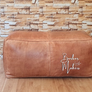 May include: A rectangular, brown leather ottoman with visible stitching and the text "Berber Makers" in white script. The ottoman is placed on a wooden floor against a brick wall.