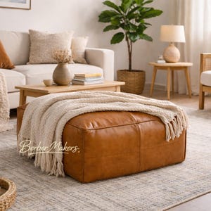 May include: A large, rectangular, brown leather ottoman with a cream-colored fringed throw blanket. The ottoman is the focal point of a living room setting, with a neutral-toned rug and a beige sofa in the background.