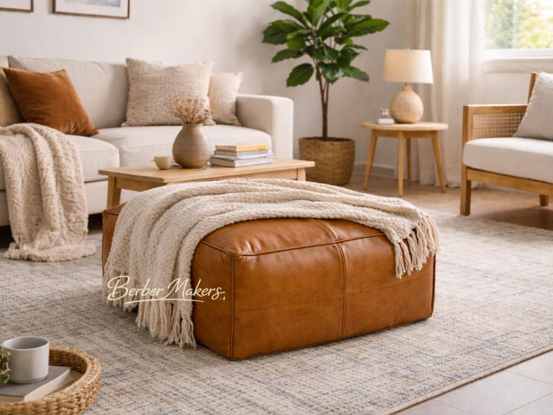 May include: A large, rectangular, brown leather ottoman with a cream-colored fringed throw blanket. The ottoman is the focal point of a living room setting, with a neutral-toned rug and a beige sofa in the background.