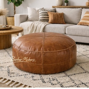 Pode incluir: Um pufe redondo de couro castanho com detalhes de costura. O pufe é o ponto focal de uma sala de estar, com um sofá branco, um tapete estampado e uma mesa de centro de madeira ao fundo. O pufe tem aproximadamente 91 cm de diâmetro.