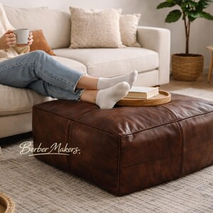 May include: A large, square, brown leather ottoman. The ottoman is in a living room setting with a person relaxing on a cream-colored sofa. A wooden tray with a book sits on top of the ottoman. The text "Berber Makers" is visible.