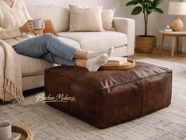 May include: A large, square, brown leather ottoman. The ottoman is in a living room setting with a person relaxing on a cream-colored sofa. A wooden tray with a book sits on top of the ottoman. The text "Berber Makers" is visible.