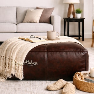 May include: A brown leather ottoman with a cream-colored knitted throw blanket and fringe. A book, eyeglasses, and a mug sit on top. The scene includes a sofa, side table, and a woven chair, creating a cozy living room setting.