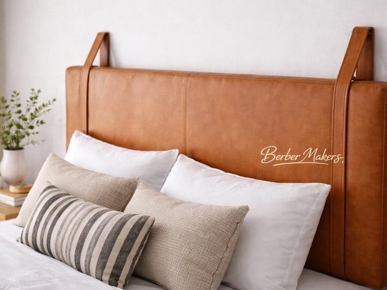 May include: A tan leather headboard with hanging straps, complemented by white and patterned pillows. The headboard is rectangular and positioned above a bed. The pillows provide texture and contrast to the bed's design.