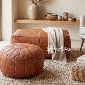 May include: A brown leather ottoman and a rectangular leather bench with a white blanket draped over it. A round, stitched ottoman sits in front. The scene is set on a patterned rug, with a shelf of decor in the background.