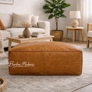 May include: A large, rectangular, brown leather ottoman with visible stitching. The ottoman is the focal point of a living room setting, with a sofa, coffee table, and rug visible in the background. The text "Berber Makers" is on the ottoman.