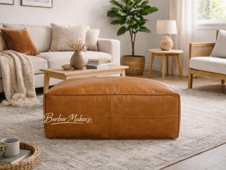 May include: A large, rectangular, brown leather ottoman with visible stitching. The ottoman is the focal point of a living room setting, with a sofa, coffee table, and rug visible in the background. The text "Berber Makers" is on the ottoman.