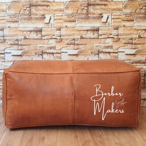 May include: A rectangular, brown leather ottoman with visible stitching. The ottoman has a soft, padded appearance and features the text "Berber Makers" in white script on the side. It sits on a wooden floor against a brick wall.