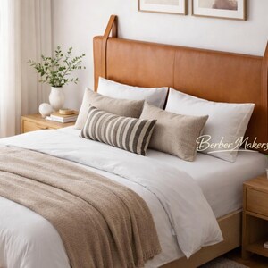 May include: A bedroom with a bed featuring a tan leather headboard. The bed is made with white bedding, a beige throw, and decorative cushions. A wooden bedside table with a lamp and plants is also visible.