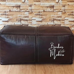 May include: A dark brown leather ottoman with white stitching details. The square-shaped pouf features a stitched design and the text "Berber Makers" in white script on the side. The background is a brick wall.