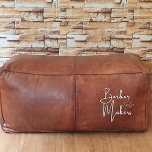 May include: A rectangular brown leather ottoman with visible stitching and a textured surface. The ottoman has the words "Berber Makers" in white script on the front. The background is a brick wall.