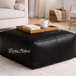 Puede incluir: Un gran puf cuadrado de cuero negro con detalles de costura. Una bandeja de madera con un libro y una taza blanca encima. El puf está en un salón con un sofá y una alfombra blancos.