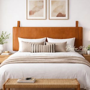 May include: A bedroom interior with a bed featuring a tan leather headboard. The bed is made with white bedding, a beige throw, and decorative pillows. Two framed abstract art pieces hang above the bed. Bedside tables and a bench complete the room.