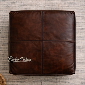 May include: A square, brown leather ottoman with a stitched cross pattern. The ottoman is a rich, dark brown color and appears to be made of high-quality leather. The ottoman is placed on a neutral-colored rug.