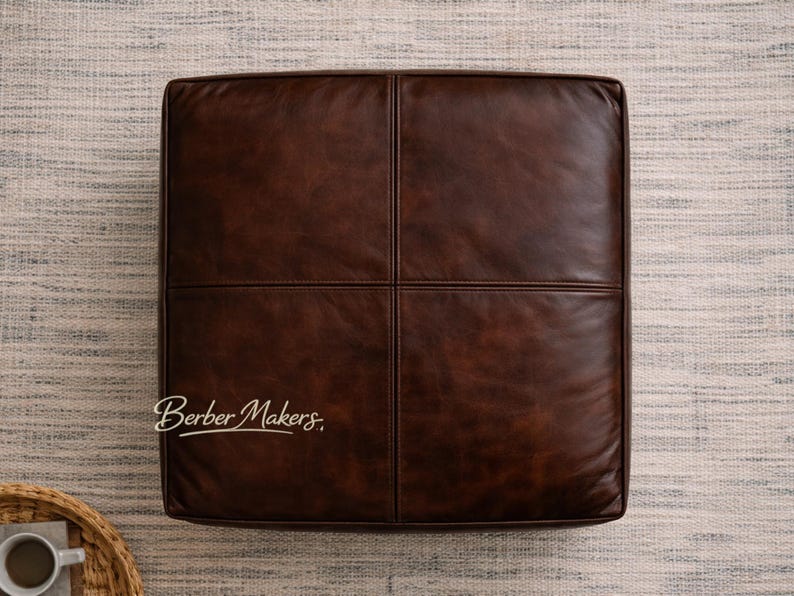 May include: A square, brown leather ottoman with a stitched cross pattern. The ottoman is a rich, dark brown color and appears to be made of high-quality leather. The ottoman is placed on a neutral-colored rug.