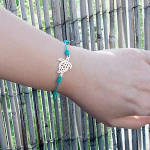 Puede incluir: Pulsera con dije de tortuga plateada y cordón turquesa. La pulsera presenta un colgante de tortuga detallado y está anudada para ajustarla. Fondo de tallos de bambú.