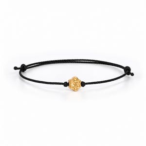May include: A black cord bracelet featuring a single gold-toned paw print charm centered on the string. The bracelet includes adjustable sliding knots on each side of the charm to secure the fit, creating a simple and functional piece of jewelry.