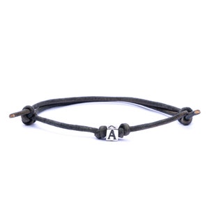 May include: A dark brown leather cord bracelet featuring a single silver-toned cube bead with the letter A engraved on its front face. The bracelet is secured with two adjustable sliding knots on either side of the central letter bead.
