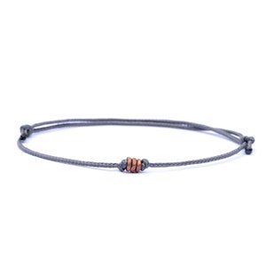 May include: A grey cord bracelet featuring three copper-coloured rings as a central design. The bracelet is knotted at both ends, set against a plain white background.