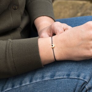 May include: A delicate bracelet featuring a thin black cord and a small, polished gold-toned crescent moon charm centred on the wrist. The minimalist design consists of a simple string band that secures around the arm, highlighting the metallic moon.