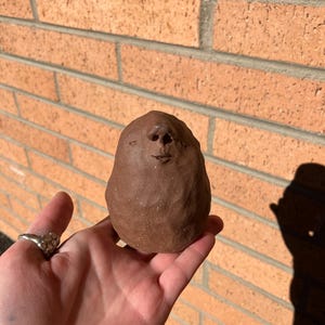 May include: A handmade, brown ceramic sculpture of a face, held in a hand. The sculpture has a rounded shape with facial features. The background is a brick wall.