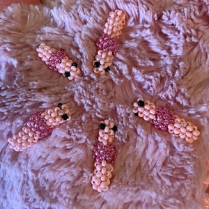 Puede incluir: Seis pinzas para el pelo con cuentas en tonos rosa, melocotón y negro están dispuestas sobre una superficie rosa esponjosa. Las pinzas están hechas de pequeñas cuentas, con una sección rosa más oscura en el medio y cuentas negras en los extremos.