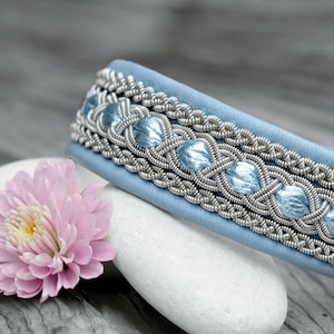 Op de afbeelding: Een lichtblauwe armband met ingewikkeld zilverdraadwerk en lichtblauwe accenten. De armband ligt op een witte steen naast een roze bloem. Het ontwerp heeft geweven patronen en een zacht, getextureerd uiterlijk.