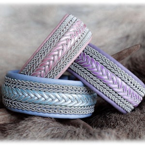 Op de afbeelding: Drie handgemaakte armbanden in roze, paars en blauw. Elke armband heeft een gevlochten ontwerp met zilverkleurige metalen accenten. De armbanden worden gepresenteerd op een getextureerd, neutraal getint oppervlak, dat hun details benadrukt.