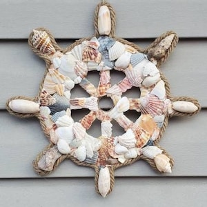 Pode incluir: Uma decoração de parede com tema náutico em forma de leme. O leme é feito de conchas em várias cores, incluindo branco, rosa e bege, e é contornado com corda. Ele é pendurado por um laço de corda.