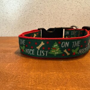 Puede incluir: Un collar verde para perros con ribete rojo, con el texto "THE NICE LIST" y gr&aacute;ficos festivos de &aacute;rboles de Navidad y huesos. El collar tiene una hebilla negra y un lazo met&aacute;lico para sujetar la correa.