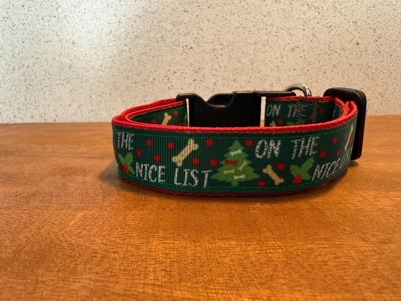 Puede incluir: Un collar verde para perros con ribete rojo, con el texto "THE NICE LIST" y gr&aacute;ficos festivos de &aacute;rboles de Navidad y huesos. El collar tiene una hebilla negra y un lazo met&aacute;lico para sujetar la correa.