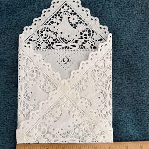 May include: A white, laser-cut paper envelope with intricate floral and crown designs. The envelope is open, revealing a glimpse of the interior. A wooden ruler is placed next to the envelope, showing measurements in inches.