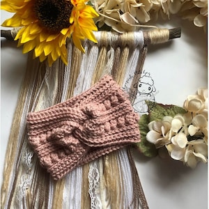 May include: A pink crocheted headband with a cable knit pattern and a textured design. The headband is displayed against a backdrop of yarn strands and dried flowers.