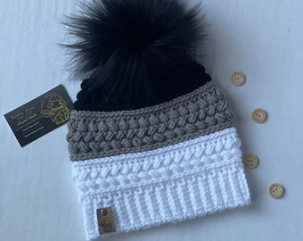 May include: A white, gray, and black crocheted beanie with a black faux fur pom pom. The beanie has a white crocheted band with a gray crocheted band above it. The beanie has a black crocheted band at the top with a black faux fur pom pom.
