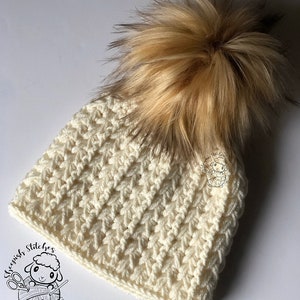 May include: A white crocheted beanie with a brown and blonde faux fur pom pom. The beanie has a textured stitch pattern and a folded brim.