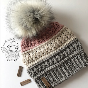 以下が含まれることがあります： ふわふわの白いポンポンが付いたグレー、白、ピンクのクロシェハット。ハットはテクスチャのあるステッチパターンで、"Sheepish Stitches"と書かれた木製タグで飾られています。