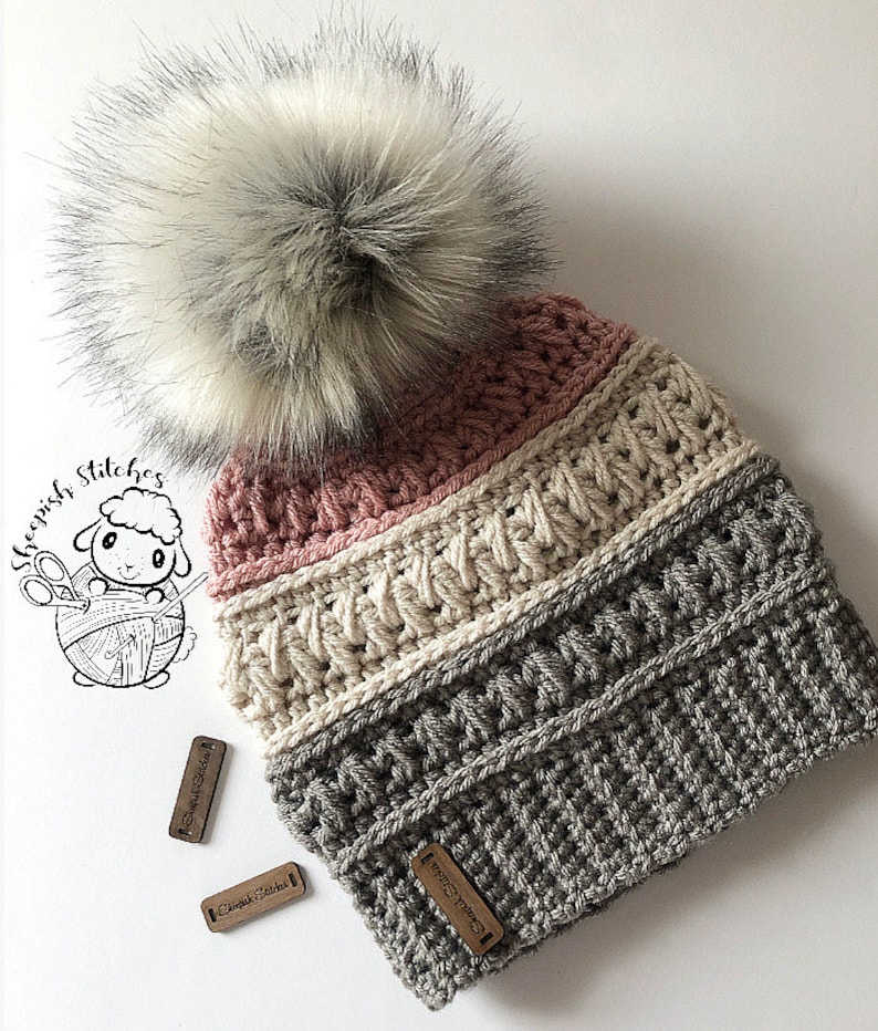 May include: A gray, white, and pink crocheted beanie with a white faux fur pom pom. The beanie has a textured stitch pattern and is decorated with wooden tags that say "Sheepish Stitches".