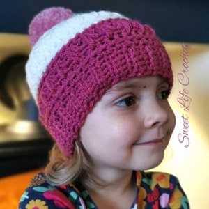 May include: A pink and white crocheted hat with a fluffy pom pom on top. The hat is worn by a young child with blonde hair.