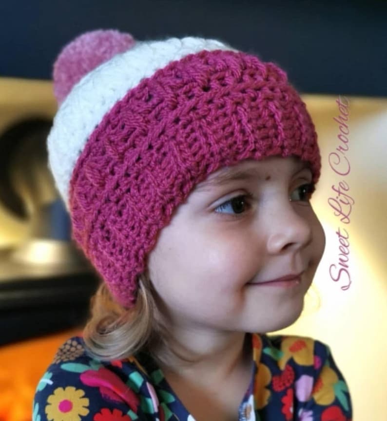 May include: A pink and white crocheted hat with a fluffy pom pom on top. The hat is worn by a young child with blonde hair.