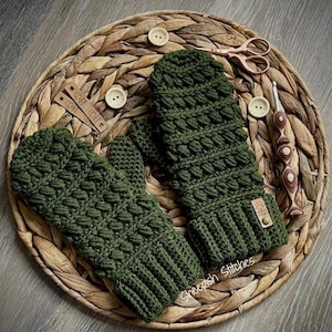 May include: A pair of olive green crochet mittens with a textured stitch pattern. The mittens are displayed on a woven basket, alongside wooden buttons, scissors, and a crochet hook. A small leather tag is attached to one mitten.