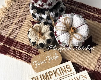 Può includere: Quattro zucche all'uncinetto in vari colori e motivi, tra cui bianco, grigio, nero e bordeaux, con un cartello di legno che recita "Farm Fresh Pumpkins Pick Your Own".