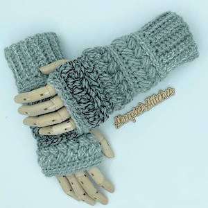 May include: A pair of grey crocheted fingerless gloves with a cable knit pattern. The gloves are on a wooden hand model. The image includes the text "sheepishstitches".