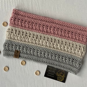 Puede incluir: Un calentador de cuello de punto en rosa, crema y gris con un patrón de punto de cable tejido. El calentador de cuello tiene una etiqueta de cuero marrón con el texto "Bobbie Anne" y una pequeña ilustración de oveja.