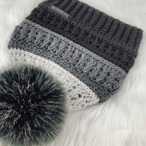 May include: A gray, white, and black crocheted beanie with a fluffy pom pom. The beanie has a label that says "Love Stitches ATL".
