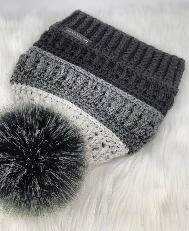 May include: A gray, white, and black crocheted beanie with a fluffy pom pom. The beanie has a label that says "Love Stitches ATL".