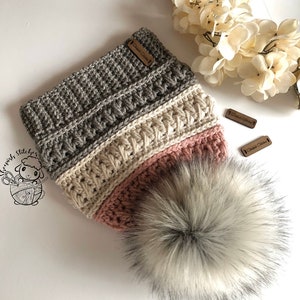 Peut inclure: Un bonnet en crochet gris, crème et rose avec un pompon blanc moelleux. Le bonnet porte une étiquette en cuir marron qui dit "Crochet Stitches".
